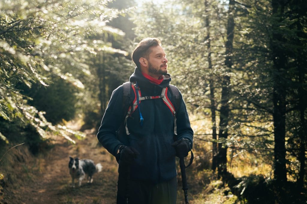 Serious man hiking with backpack and trekking pole in a dense forest during daylight, exploring nature and outdoor adventure.