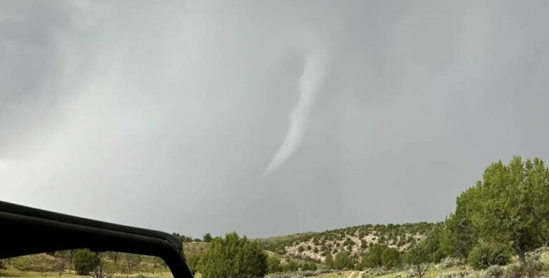 A tornado forming over a rural landscape with trees and hills, under a cloudy sky, illustrating severe weather conditions relevant to storm preparedness.
