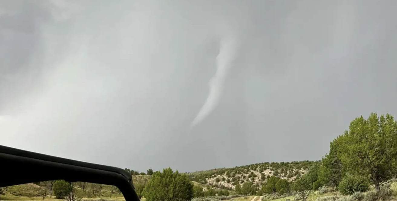A tornado forming over a rural landscape with trees and hills, under a cloudy sky, illustrating severe weather conditions relevant to storm preparedness.